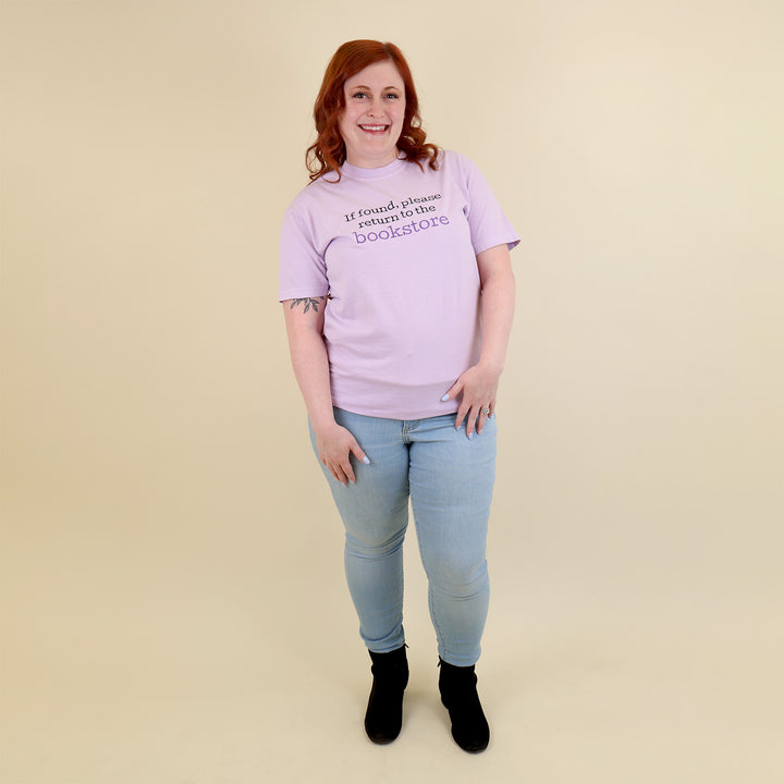 A woman wearing a light purple t-shirt with 'If found, please return to the bookstore' embroidered on it, standing on a beige background.