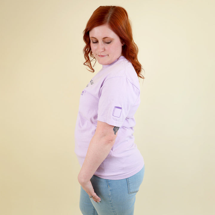 A woman wearing a light purple t-shirt with 'If found, please return to the bookstore' embroidered on it and a book embroidered on the sleeve, standing on a beige background.