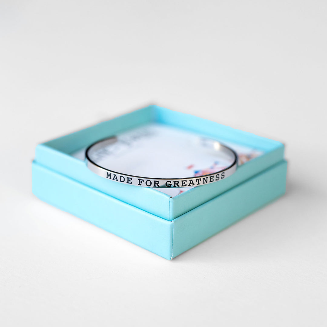 a silver cuff bracelet embossed with the words "made for greatness" sits on the edge of its box.