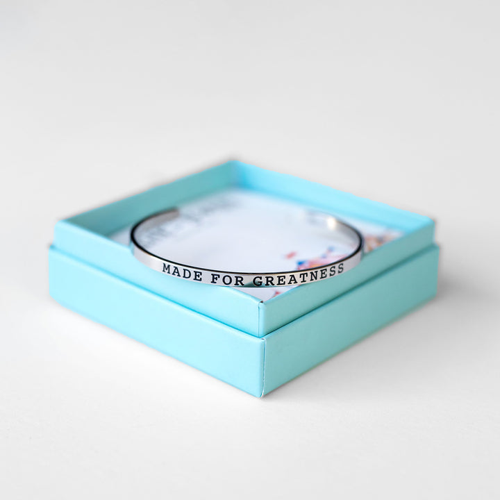 a silver cuff bracelet embossed with the words "made for greatness" sits on the edge of its box.