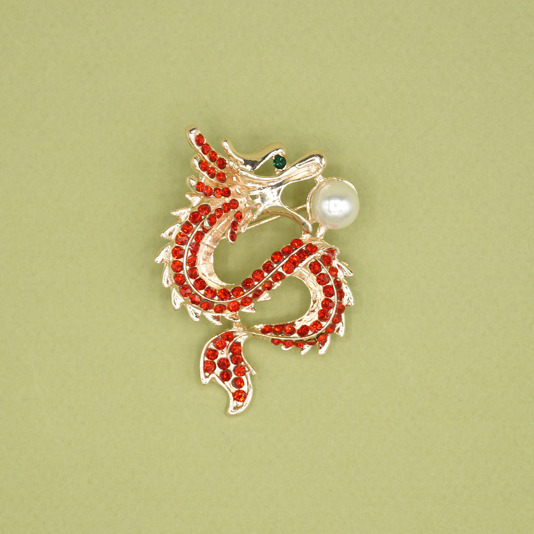 A gold dragon-shaped brooch with red sequins along the body, a green jewel eye, and a pearl in its mouth.