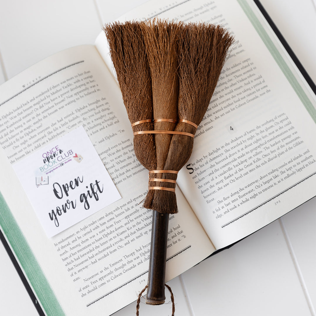 A miniature broom lays across an open book next to an open your gift sticky note.