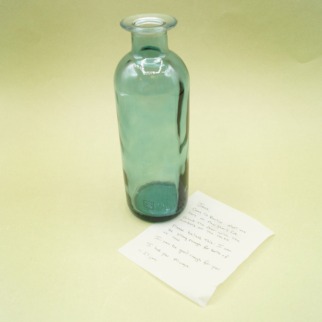 A green-blue bottle vase next to a white piece of paper with handwriting on it.