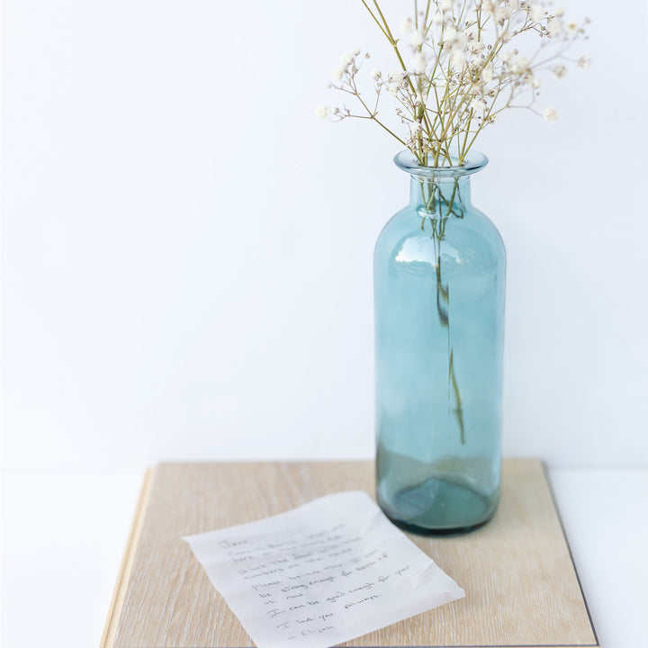 A green-blue bottle vase holding a small bouquet of baby's breath next to a white piece of paper with handwriting on it.