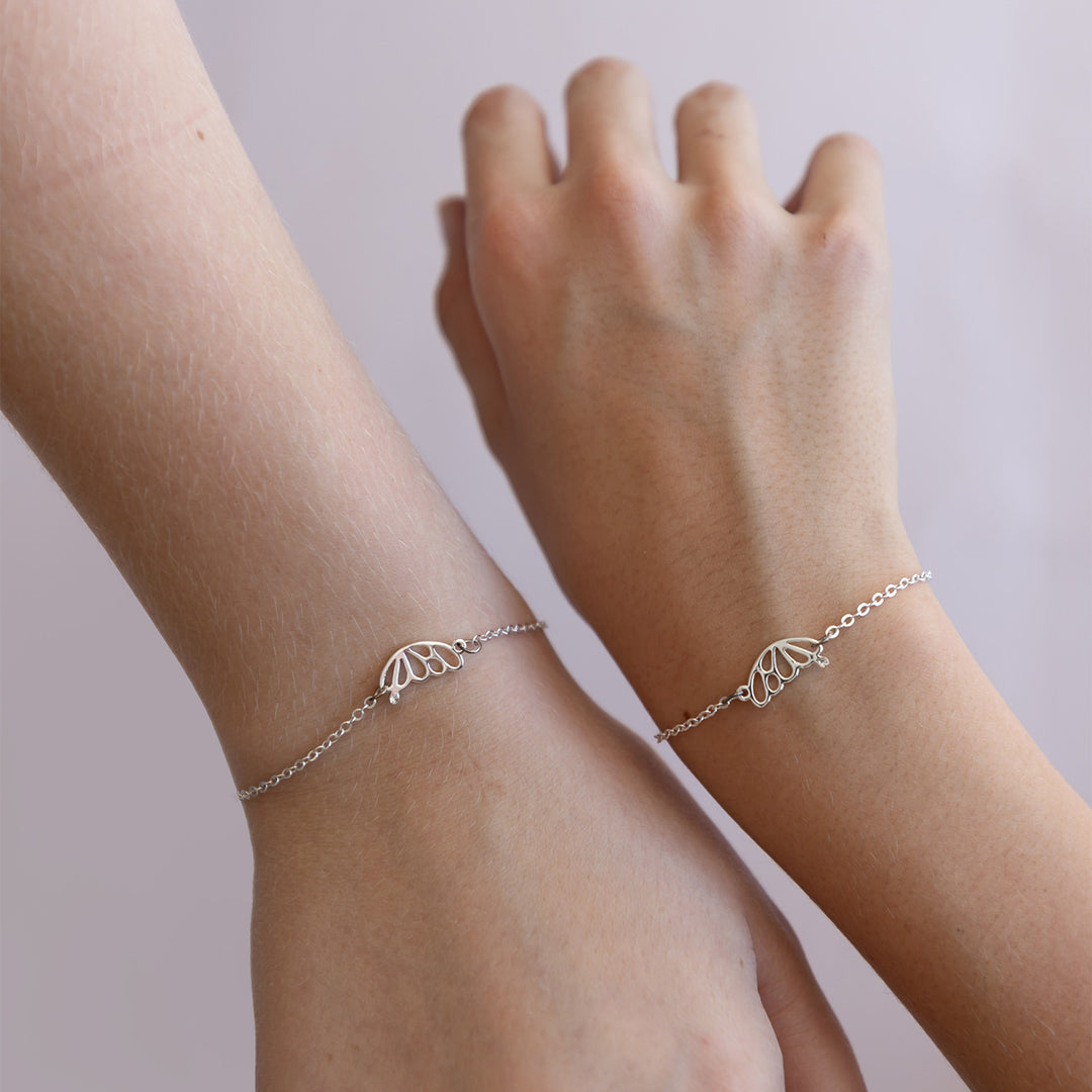 Two white arms are next to each other, each wearing a silver bracelet with a butterfly wing on each.