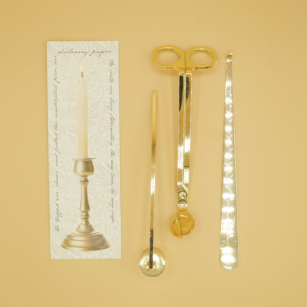 A gold candle care set - a wick trimmer, candle snuffer, and wick dipper next to a box with an image of a candlestick on it