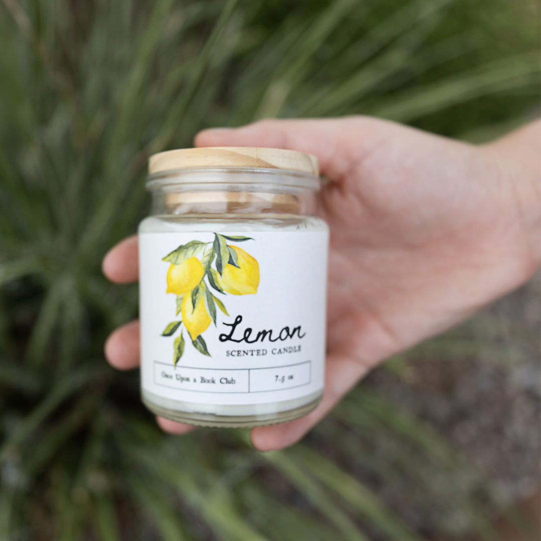 A hand holds out a glass jar candle with an image of three lemons on it. The text says: Lemon scented candle, Once Upon a Book Club, 7.5 oz.