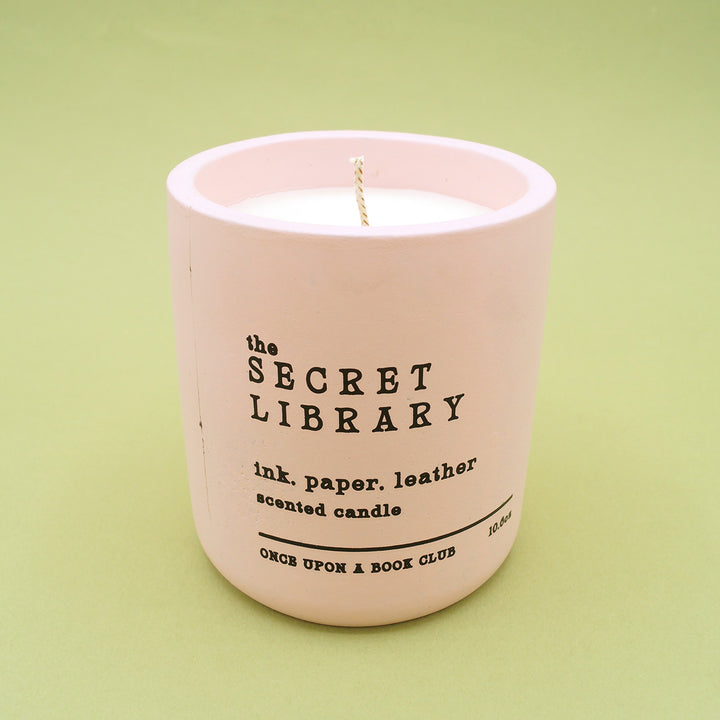 a gorgeous pink candle with vintage font "The Secret Library. ink paper leather. scented candle." on green background.