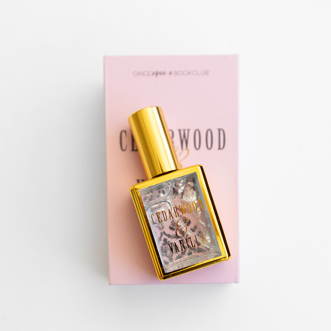 A gold perfume bottle labeled Cedarwood & Vanilla lays on a pink box.