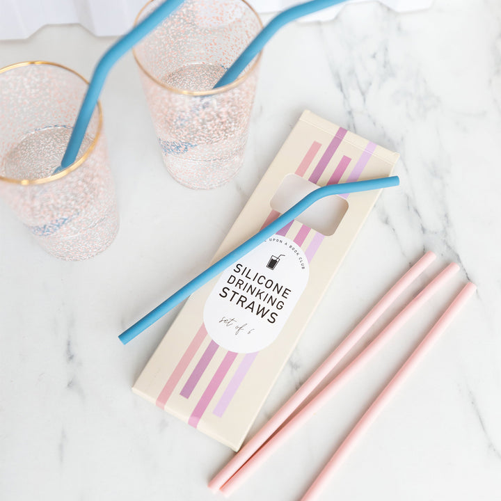 A set of 6 silicone straws sit on a countertop on their open box. The blue straws are bendy, while the pink straws are straight. Two of the blue straws are placed into drinking glasses.