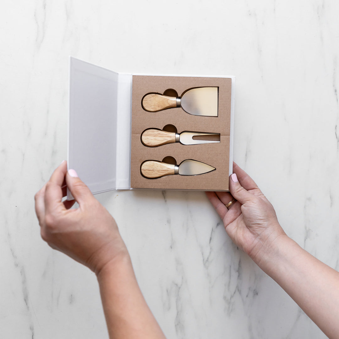 a pair of hands holds open the book-shaped box containing three cheese knives.