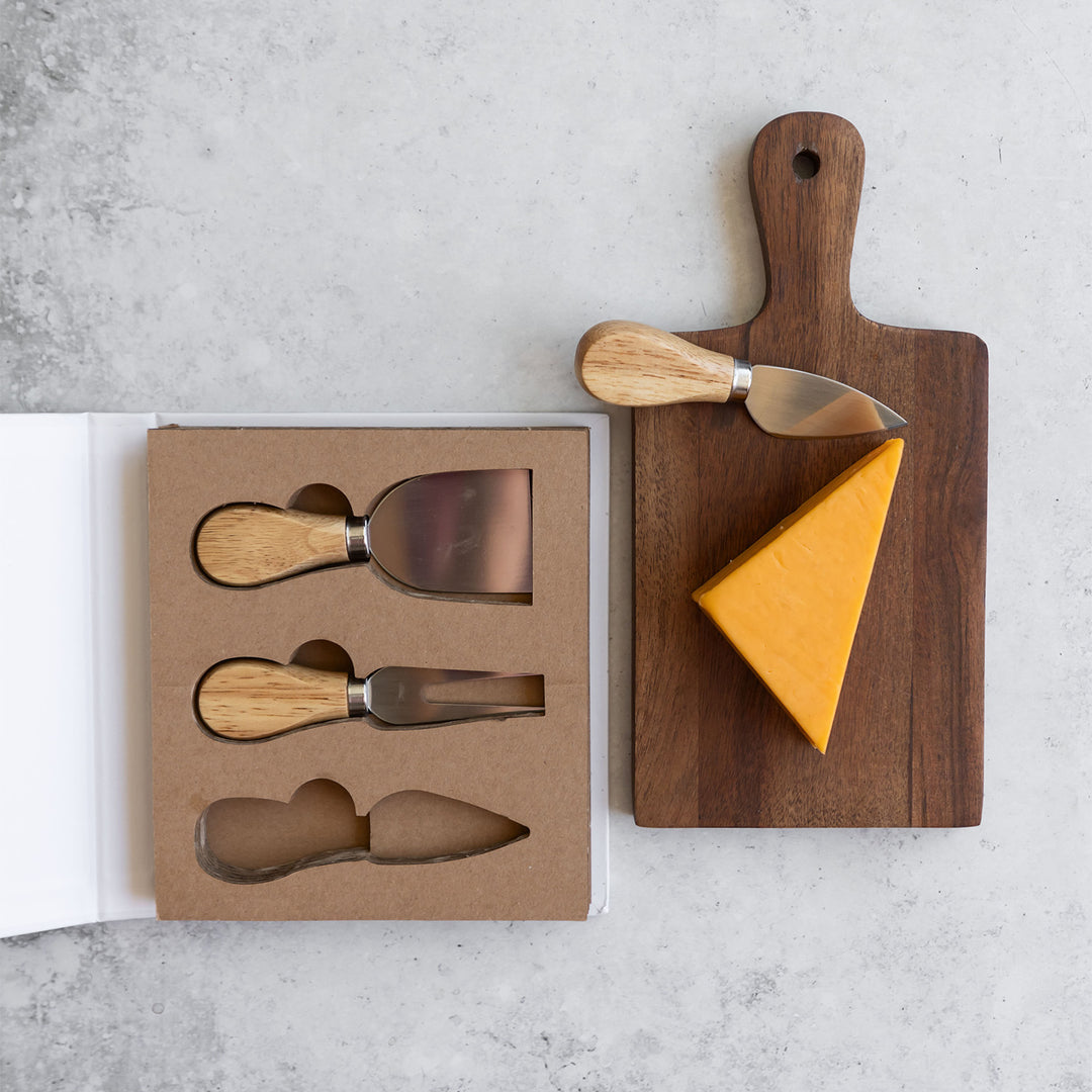 two knives sit in the book-shaped box case next to a miniature cutting board holding a wedge of cheddar and the spade cheese knife.