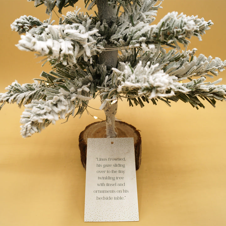 A close up of the tag attached to each miniature Christmas tree that reads "Linus frowned, his gaze sliding over to the tiny twinkling tree with tinsel and ornaments on his bedside table.