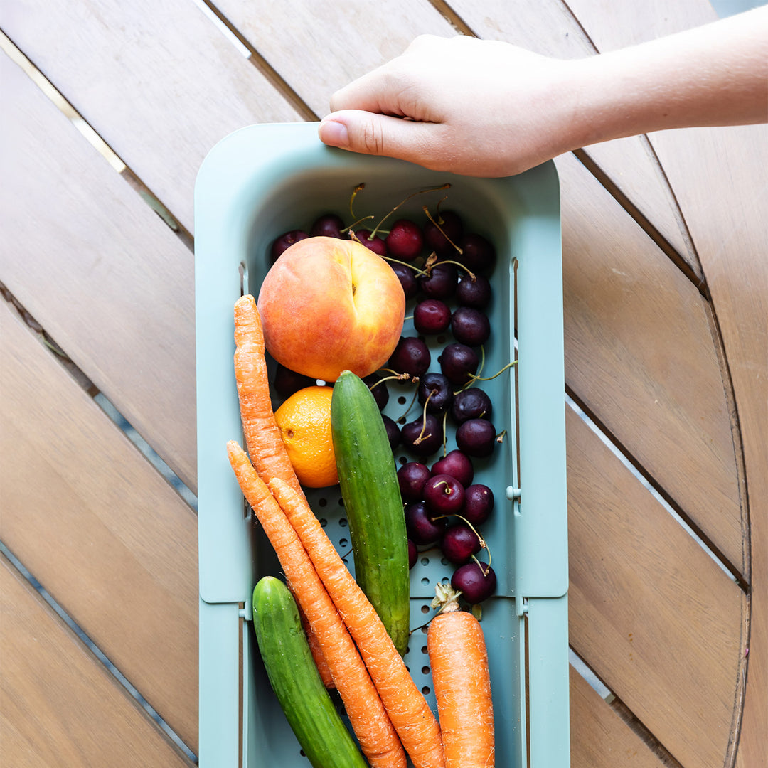 A white hand holds a pale green extendable colander filled with vegetables and fruit.