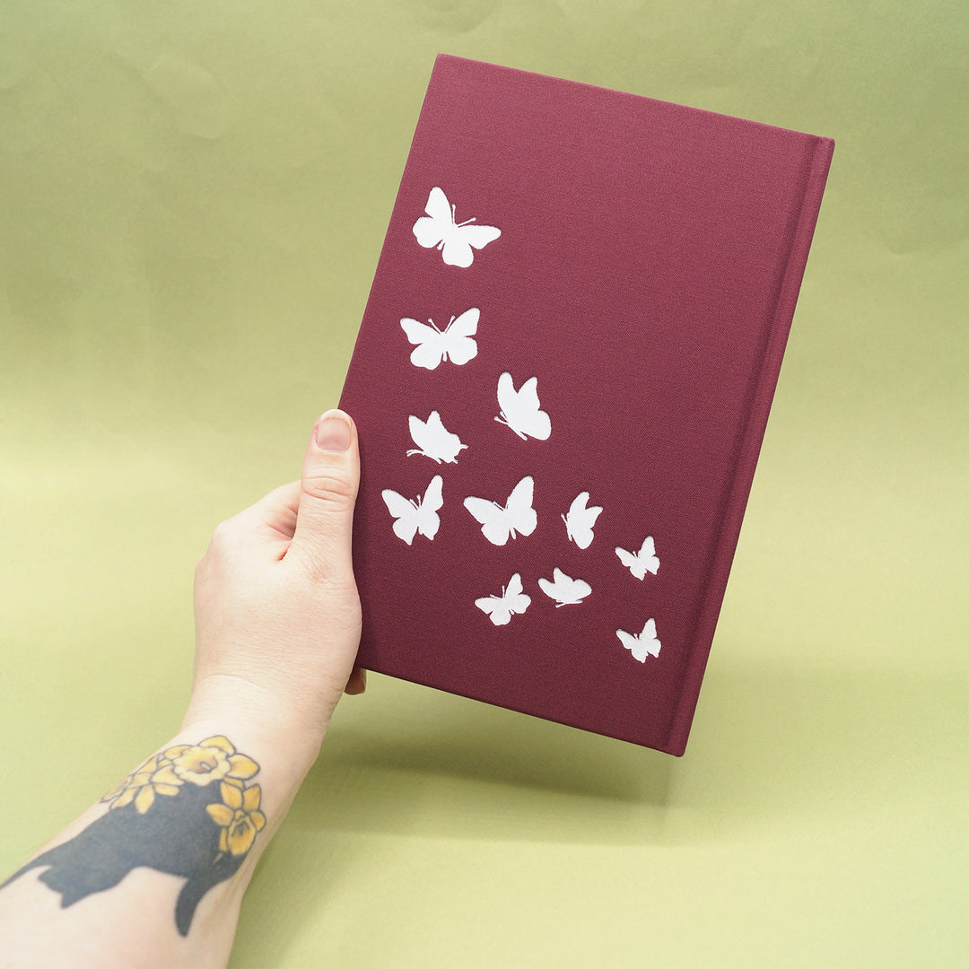 Hand holding a maroon book with white butterfly designs on a green background