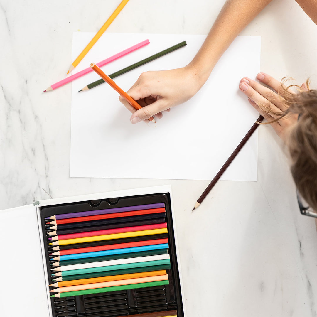 A white hand holds an orange colored pencil to a piece of paper with a few colored pencils scattered on it. Below the piece of paper is a set of colored pencils