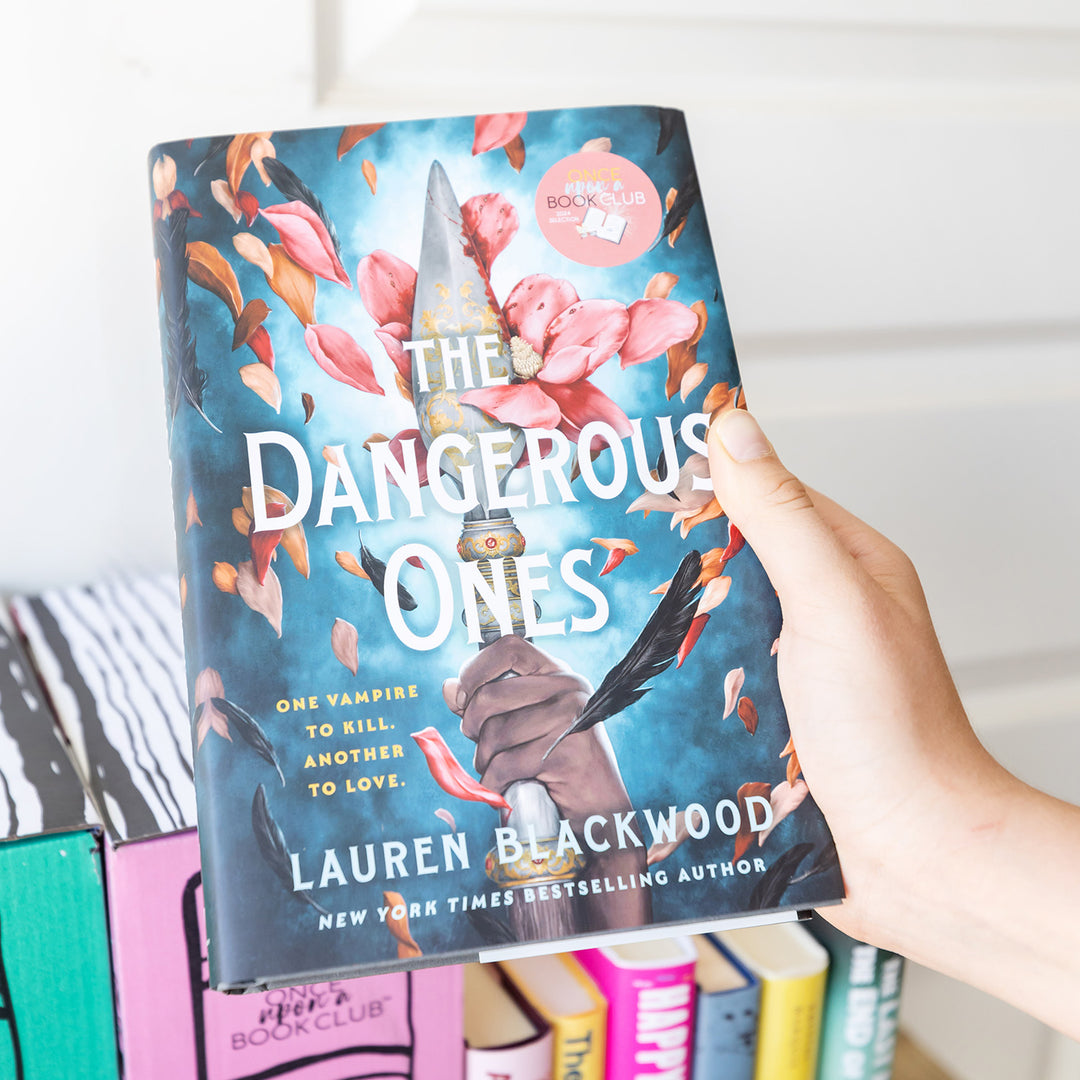 A hardcover copy of The Dangerous Ones by Lauren Blackwood is held above a stack of books.