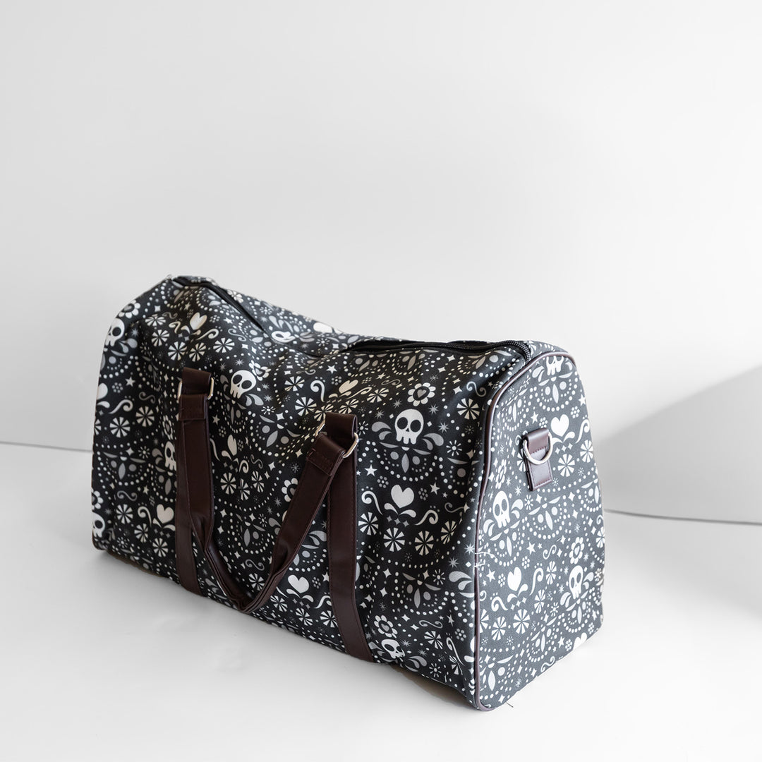 A black duffel bag with dark brown straps. The duffel bag has a pattern of white skulls, hearts, and stars on it.
