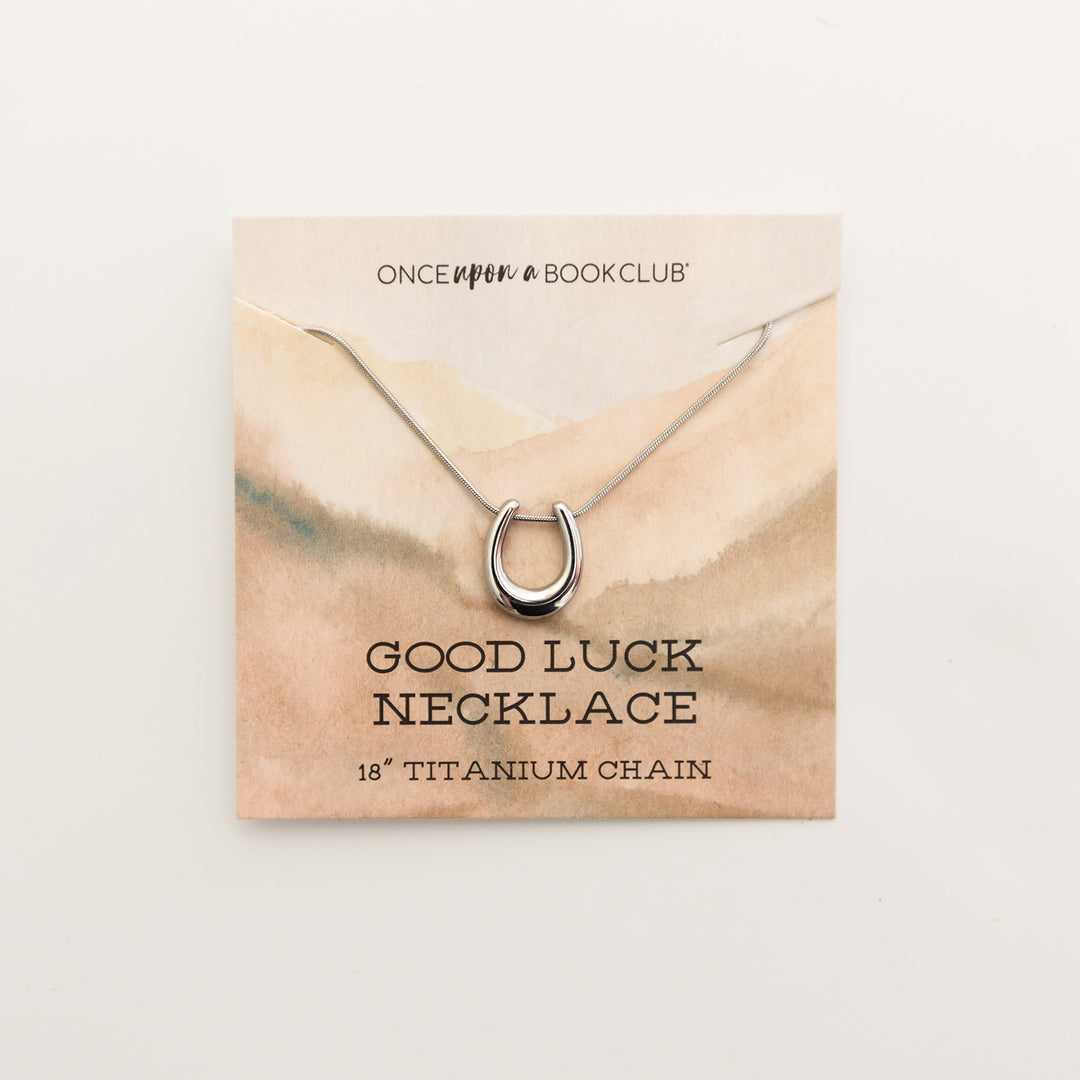 A silver horseshoe necklace on a backing card. The backing card says Once Upon a Book Club Good Luck Necklace 18" Titanium Chain.