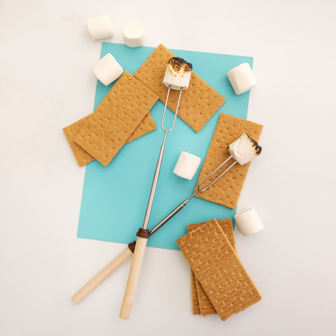 A set of silver skewers with wooden handles and burnt marshmallows on the end. The skewers lay on a blue paper with graham crackers and marshmallows surrounding them.