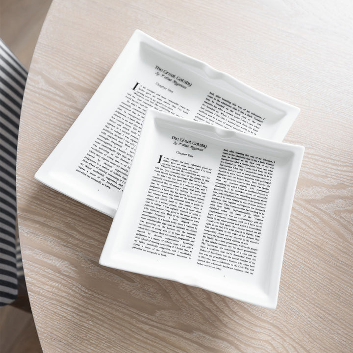 A flat lay image featuring dinner plates shaped like books inspired by The Great Gatsby by F. Scott Fitzgerald. The small plate is stacked at an angle on top of the large plate to show the size difference between the two. They sit on a wooden table.