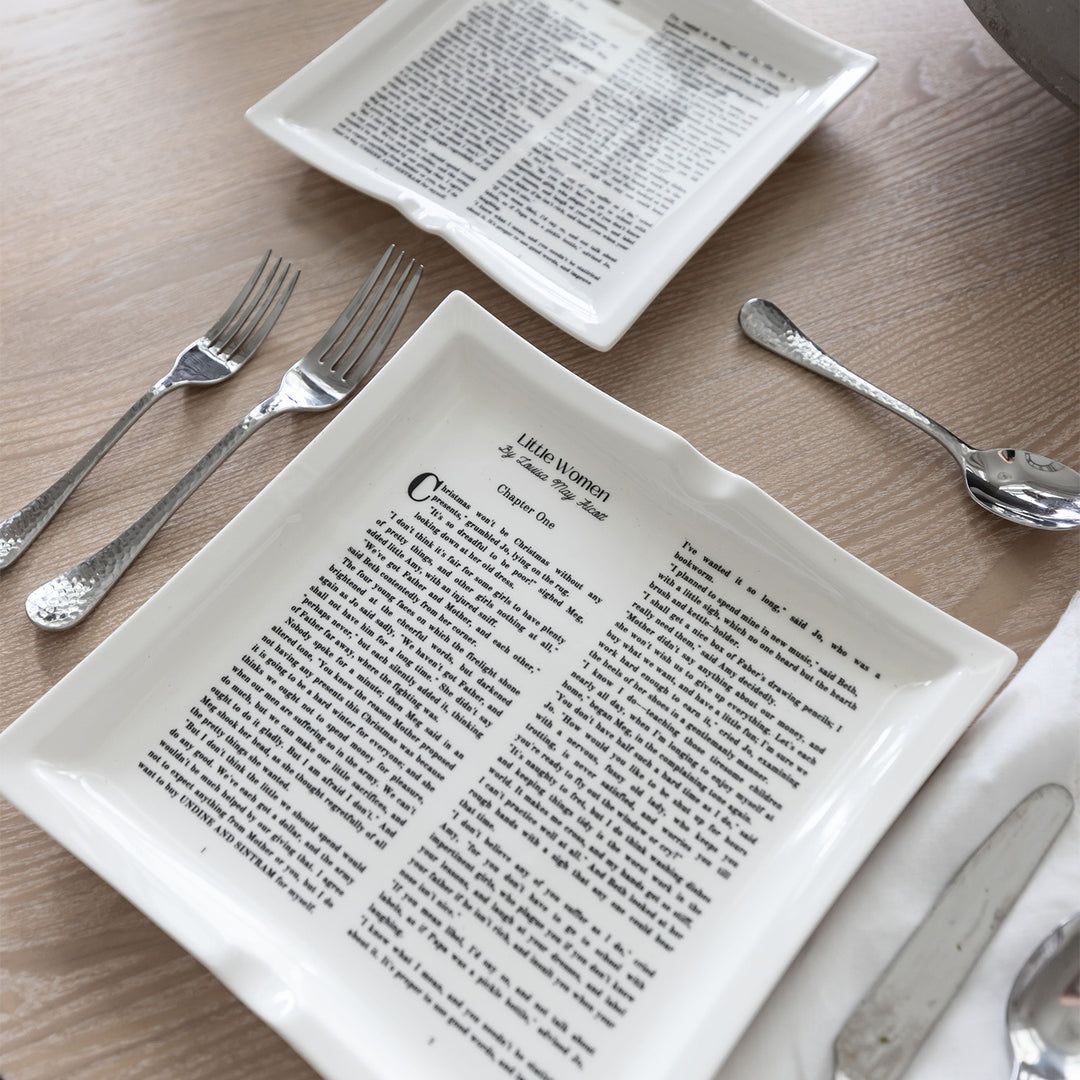 A flat lay image featuring dinner plates shaped like books inspired by Little Women by Louisa May Alcott. The large and small plate both sit on a wooden background. Silverware is placed in a standard table setting format next to the plates.