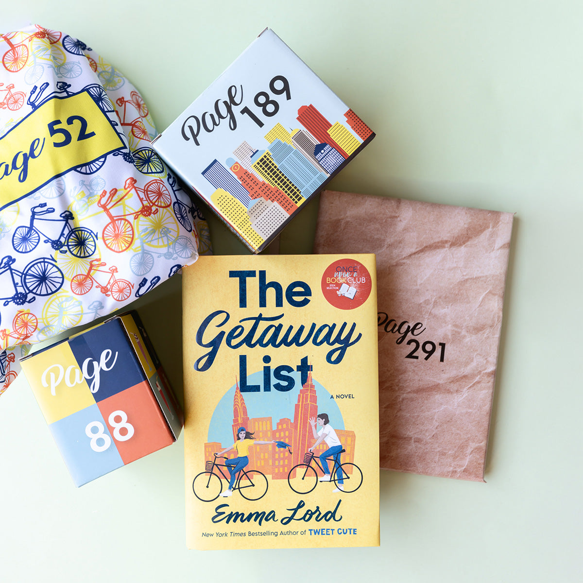 A hardcover copy of The Getaway List by Emma Lord sits on a light green background. It is surrounded by wrapped gifts labeled with page numbers in coordinating gift wrap to match the cover design of the book.