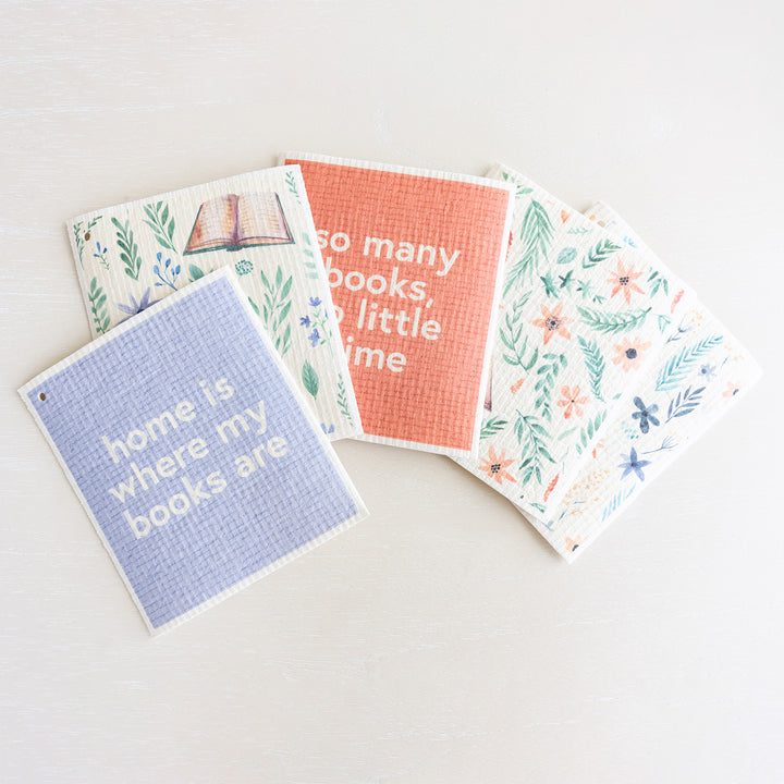 A collection of Swedish dishcloths in pastel tones featuring flowers and books. The top towel reads "home is where my books are"