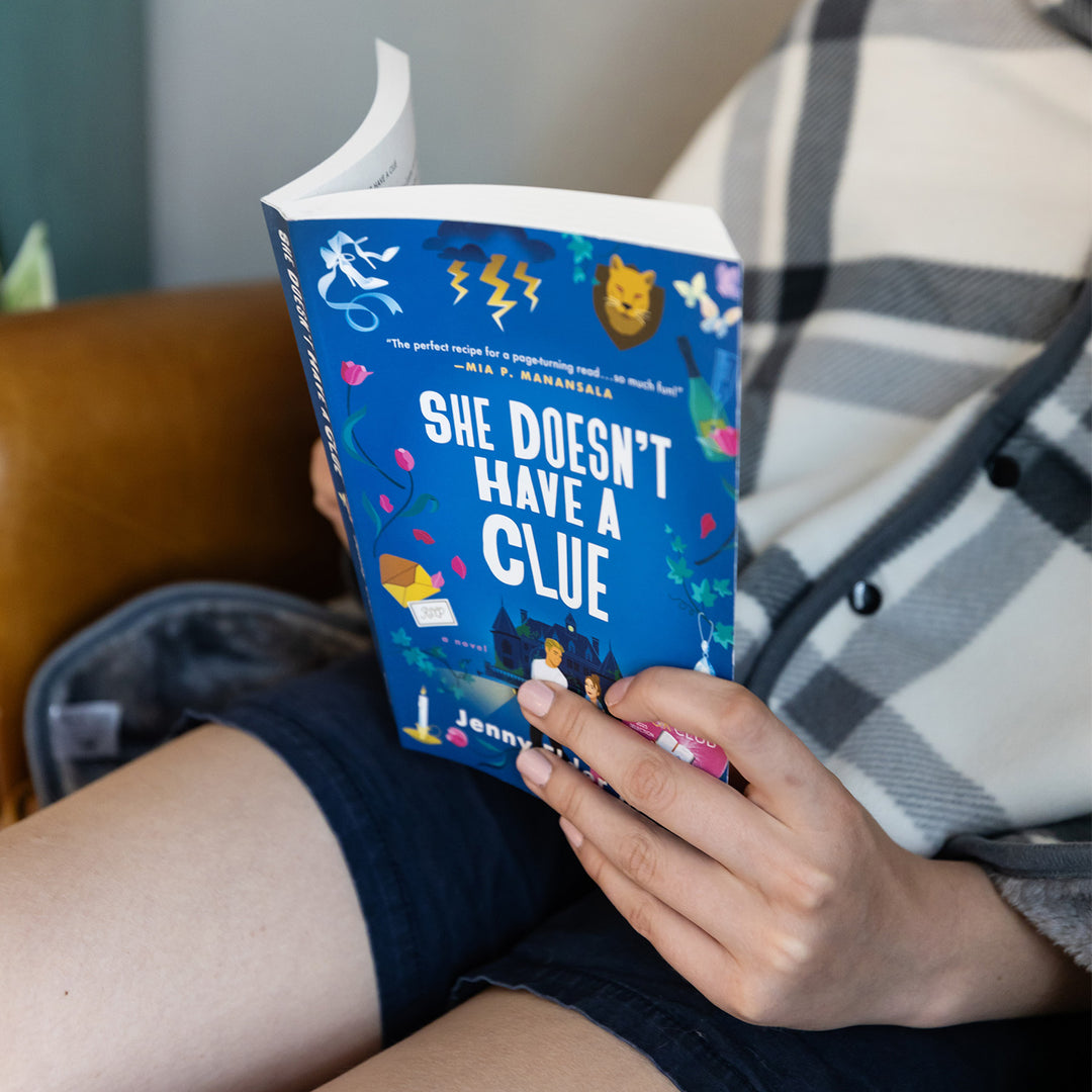 Person reading a book titled 'She Doesn't Have a Clue' by Jenny Elder Moke with colorful illustrations on the cover.