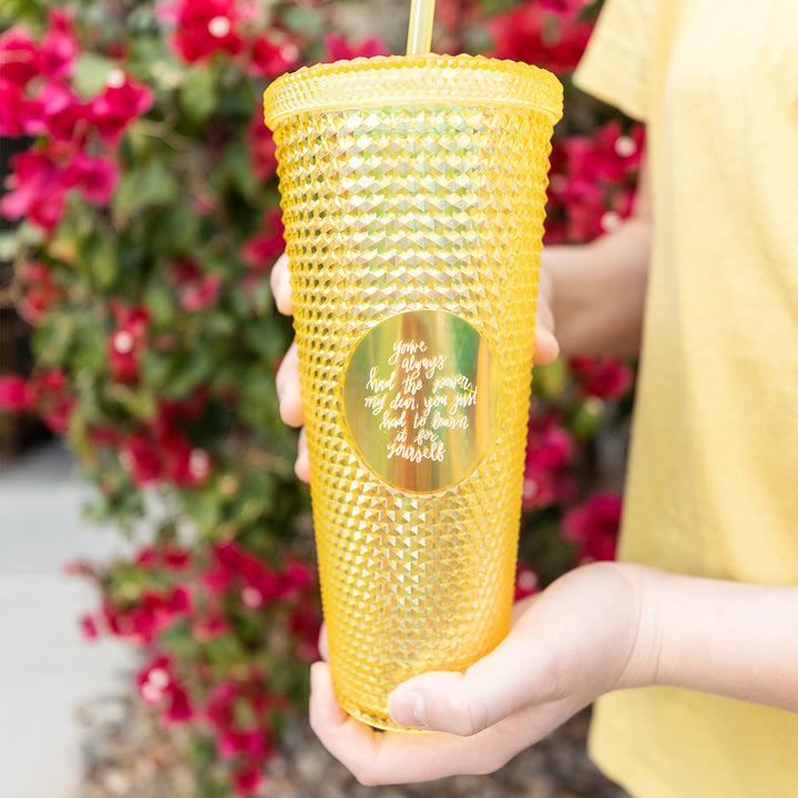 A yellow studded tumbler cup held by a pair of white hands. The cup is etched with the quote "You've always had the power, my dear, you just had to learn it for yourself."