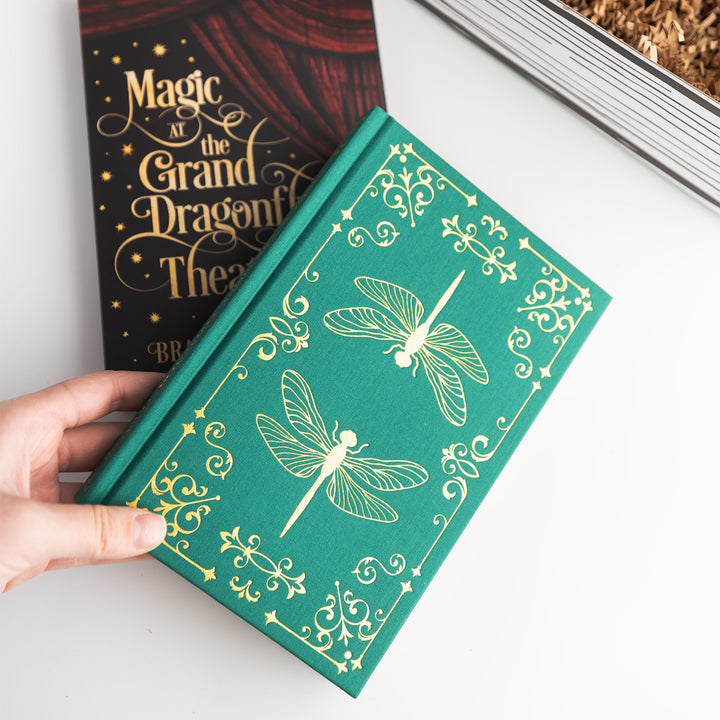 Green fabric hard case with dragonfly design held by a hand, with the dust jacket for 'Magic at the Grand Dragonfly Theatre' by Brandie June is visible beneath it.
