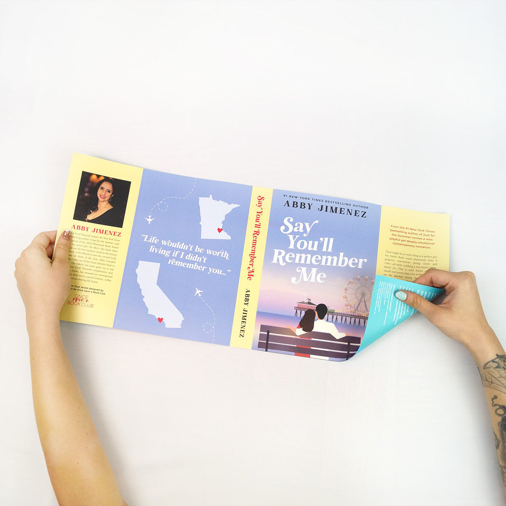 A pair of hands showing the second side of the reversible dust jacket for Say You'll Remember Me by Abby Jimenez featuring a m/f couple sitting on a bench looking at a pier with a ferris wheel. The back cover shows the outline of Minnesota and California with plane trails between the two. This is the exclusive design created by Once Upon a Book Club. 