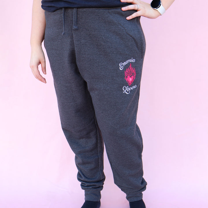 Gray sweatpants with embroidered 'Enemies to Lovers' flaming heart design on a pink background