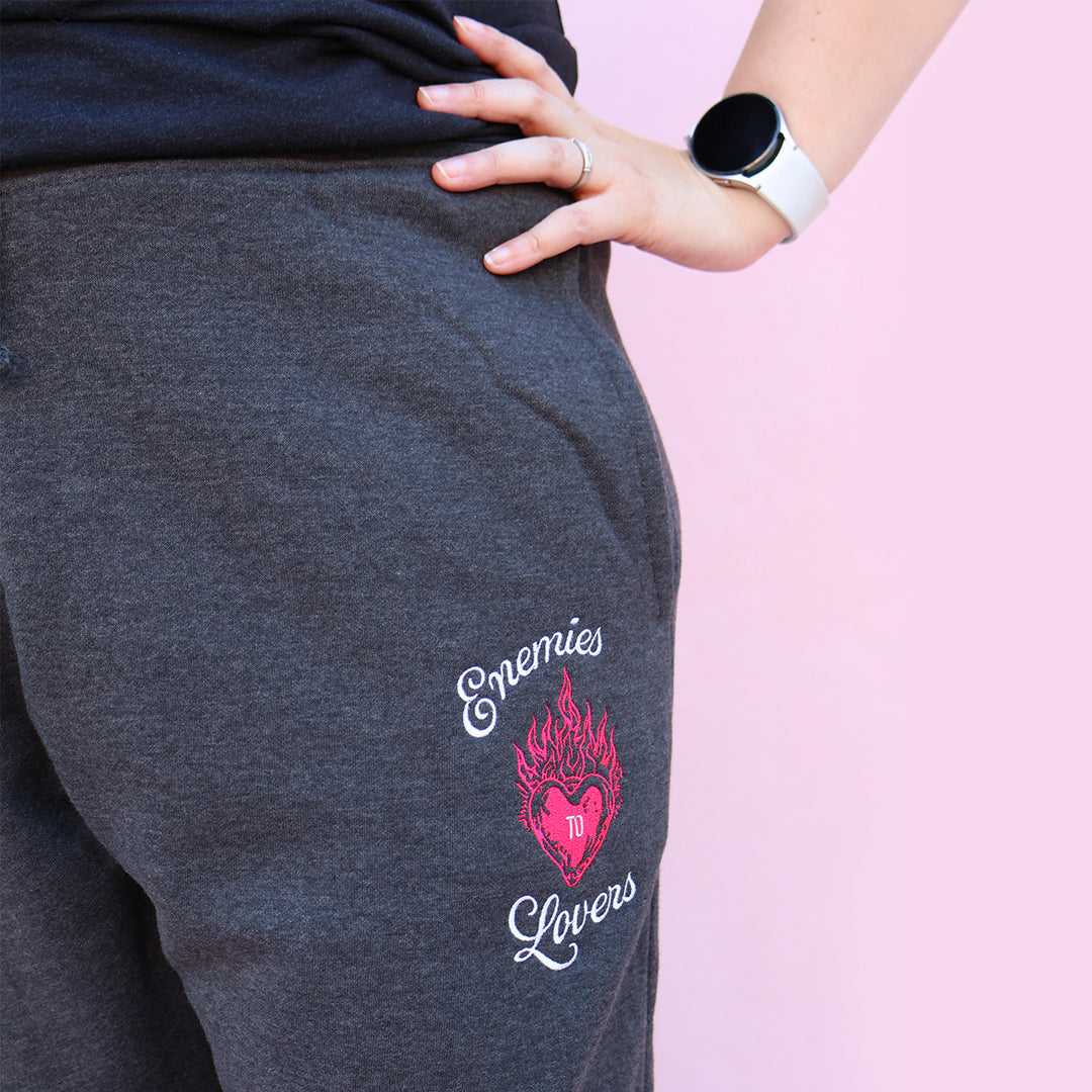 Gray sweatpants with embroidered 'Enemies to Lovers' flaming heart design on a pink background
