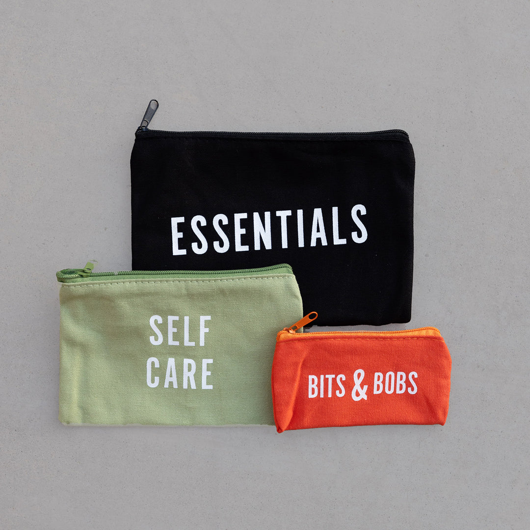 Three bags of varying sizes lay on a gray background. One bag is black and says Essentials in white letters. A slightly smaller bag is light green and says Self Care in white letters. The last and smallest bag is orange and says Bits & Bobs in white letters.