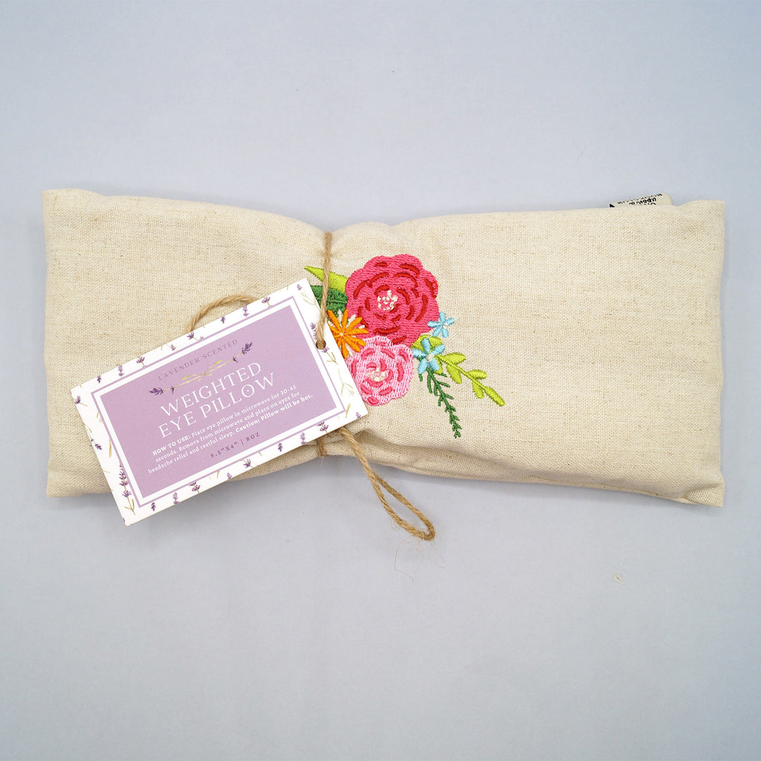 A beige weighted eye pillow with pink floral embroidery in the center.