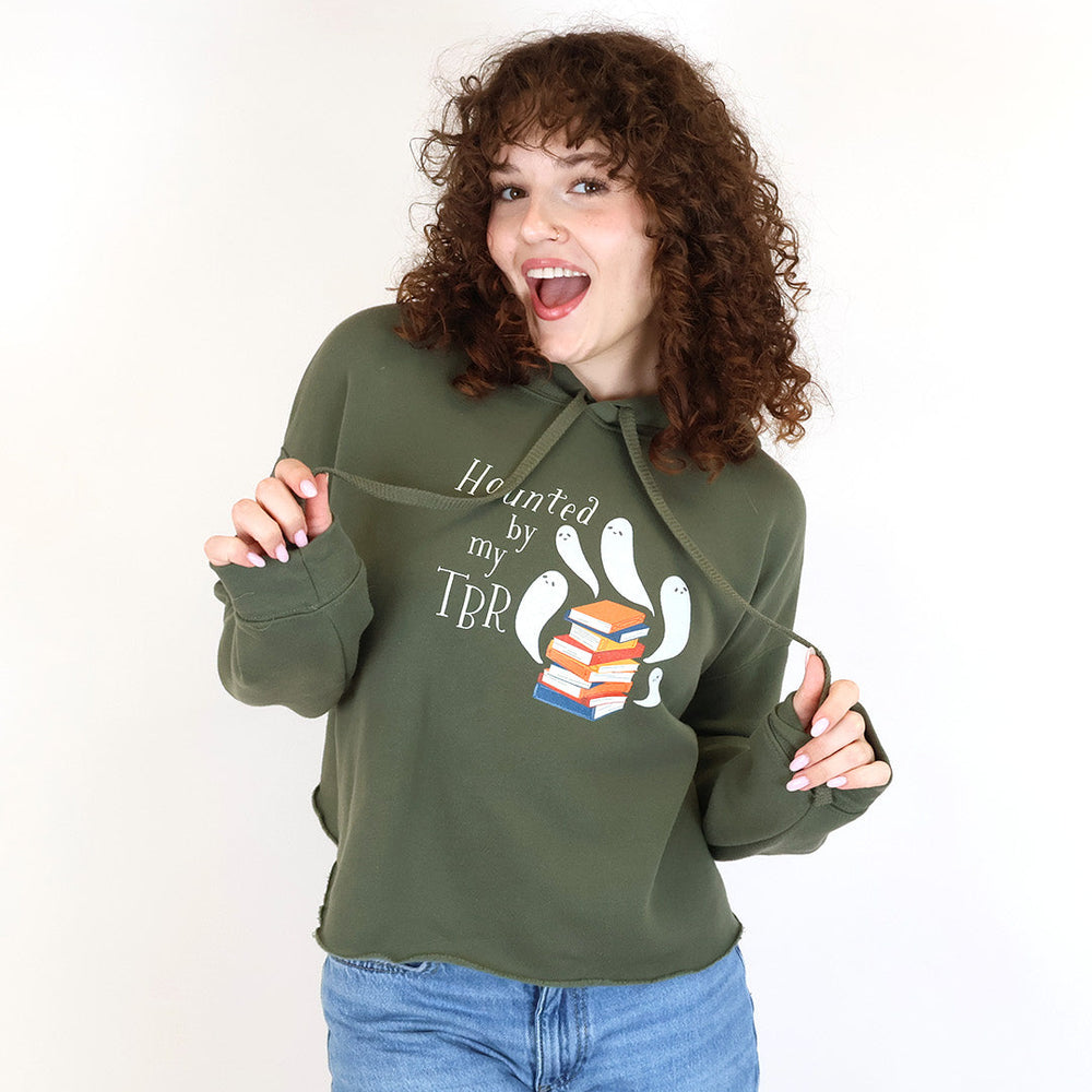 A woman with curly brown hair wearing an olive-green hoodie with a 'Haunted by my TBR' graphic and blue jeans, posing against a white studio background.