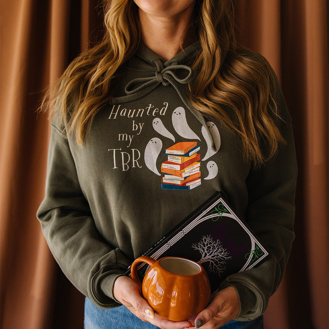 A woman wearing an olive-green hoodie with a graphic that reads 'Haunted by my TBR' and shows ghost illustrations next to a stack of books, holding an orange mug in front of a brown curtain background.