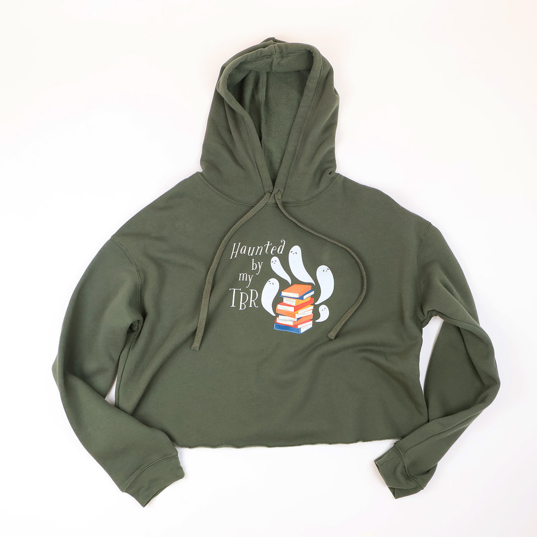 An olive-green cropped hoodie with a graphic on the chest that reads 'Haunted by my TBR' alongside cartoon ghosts and books, laid flat on a white background.