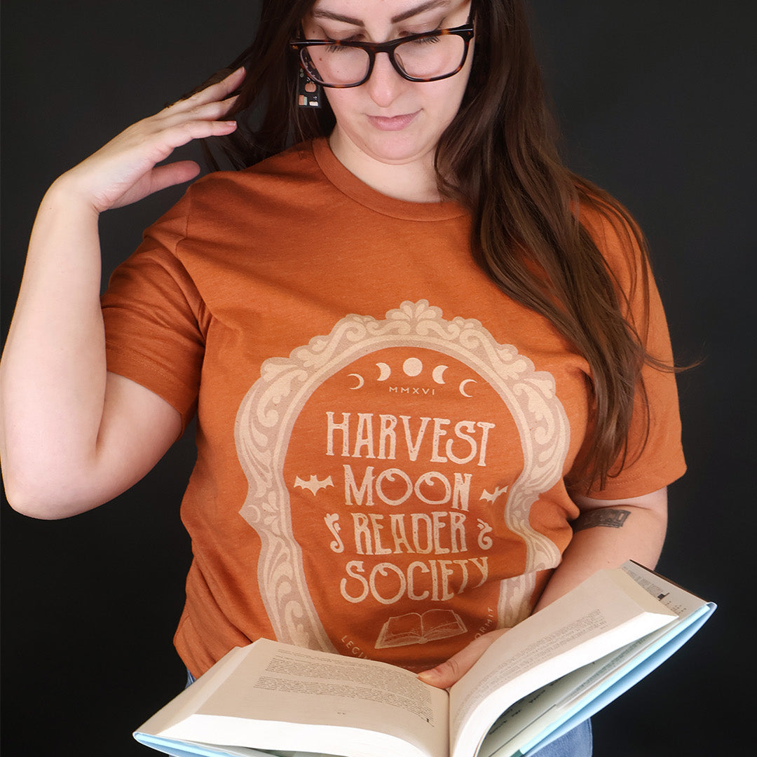 A woman wearing a rust-orange graphic t-shirt with a cream-colored 'Harvest Moon Reader Society' design, holding an open book against a dark background.