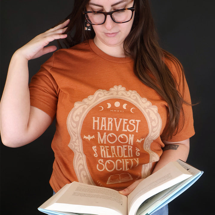A woman wearing a rust-orange graphic t-shirt with a cream-colored 'Harvest Moon Reader Society' design, holding an open book against a dark background.