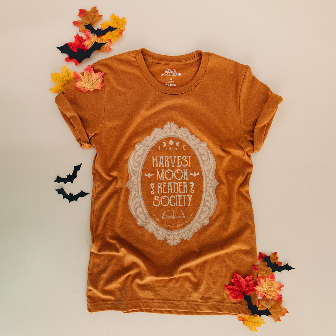 An orange-brown short-sleeve graphic T-shirt laid flat on a light beige background, surrounded by autumn leaves and small black bat cutouts. The shirt has a large cream decorative oval graphic on the front reading Harvest Moon Reader Society.