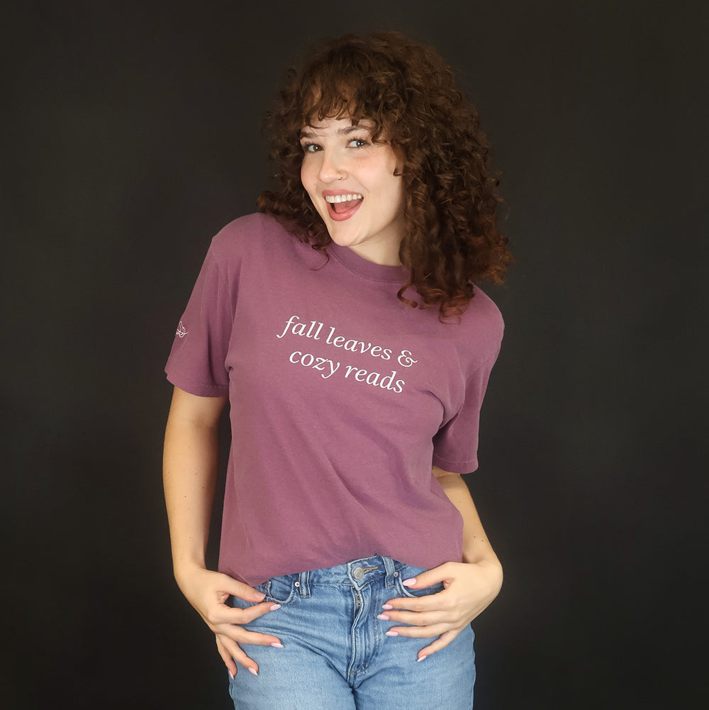 A woman with curly brown hair stands against a black studio background wearing a mauve-purple graphic t-shirt with white text that reads 'fall leaves & cozy reads' and light blue jeans.