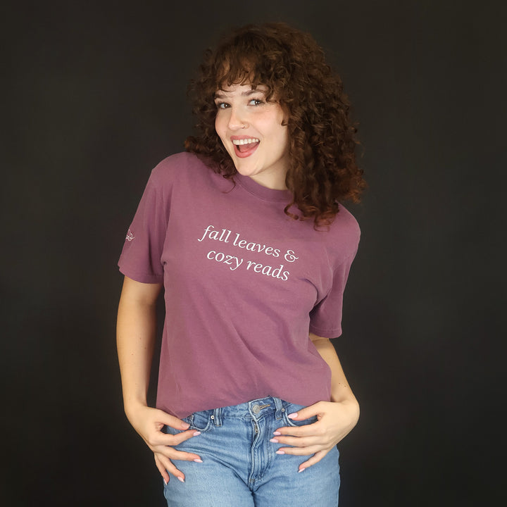 A woman with curly brown hair stands against a black studio background wearing a mauve-purple graphic t-shirt with white text that reads 'fall leaves & cozy reads' and light blue jeans.