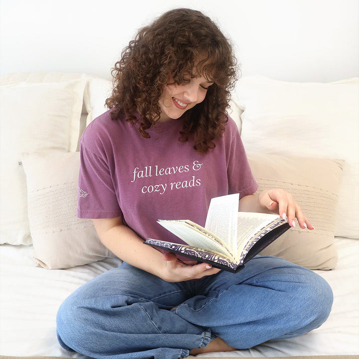 A woman with curly brown hair sits cross-legged on a bed, reading a book. She is wearing a mauve-purple graphic t-shirt with the text 'fall leaves & cozy reads' in white, paired with blue jeans.
