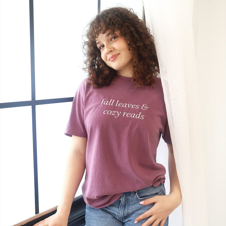 A woman with curly brown hair wearing a mauve-purple graphic t-shirt that reads 'fall leaves & cozy reads' in white script, paired with blue jeans, leaning against a white wall next to a window.