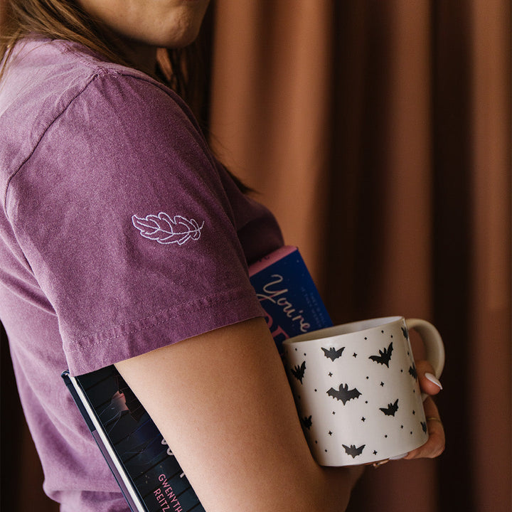 A person wearing a mauve-purple t-shirt with a white embroidered leaf design on the sleeve, holding a white ceramic mug with a black bat pattern and books under their arm, set against a brown curtain background.