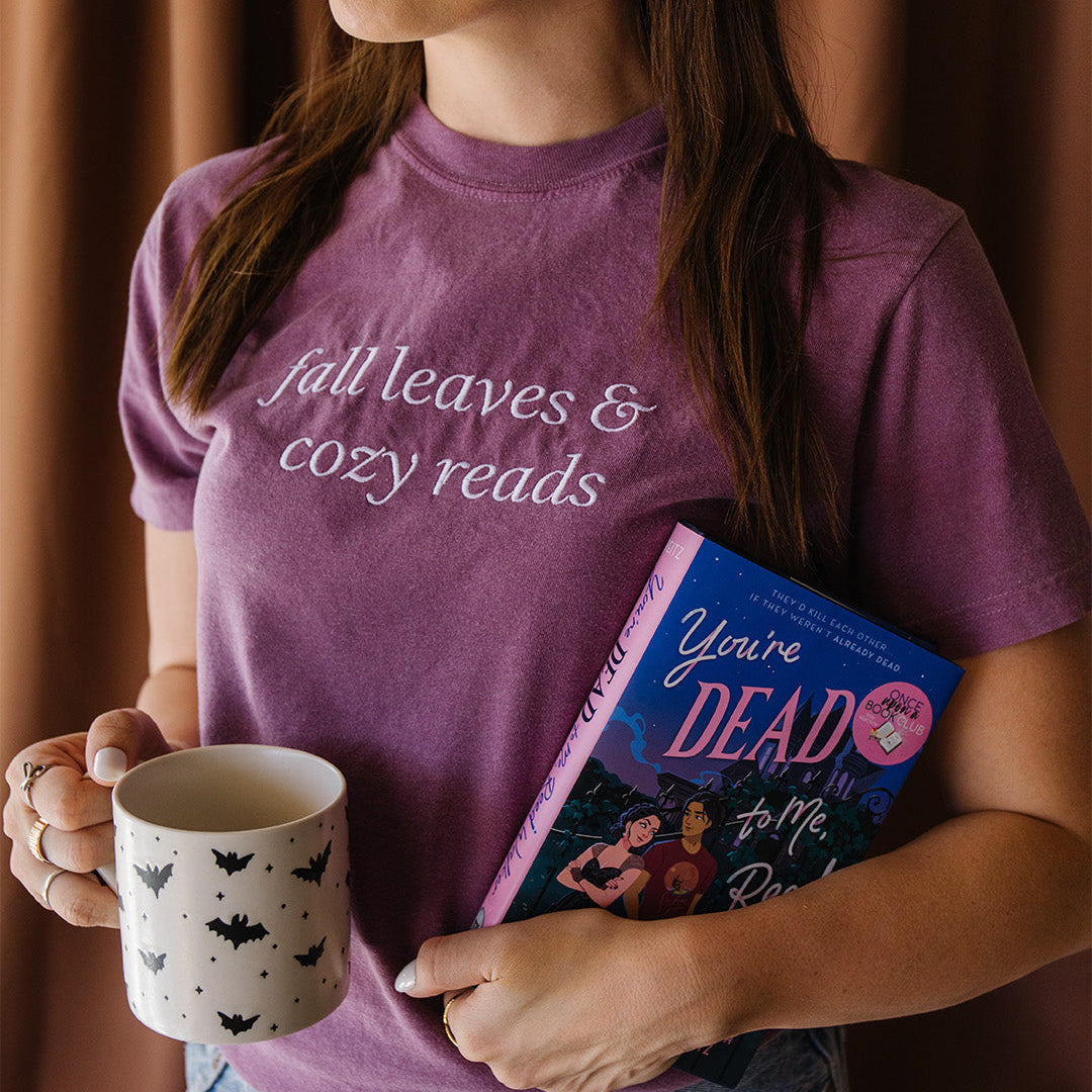 A woman wearing a purple graphic t-shirt with the text 'fall leaves & cozy reads' in white script. She is holding a white ceramic mug with black bat patterns and a book titled 'You're Dead to Me, Reed Walker'.