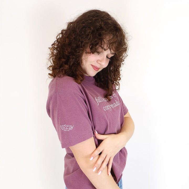 A woman with curly brown hair wearing a mauve purple graphic t-shirt and blue jeans, standing against a plain white background. There is a white embroidered leaf on the right sleeve.