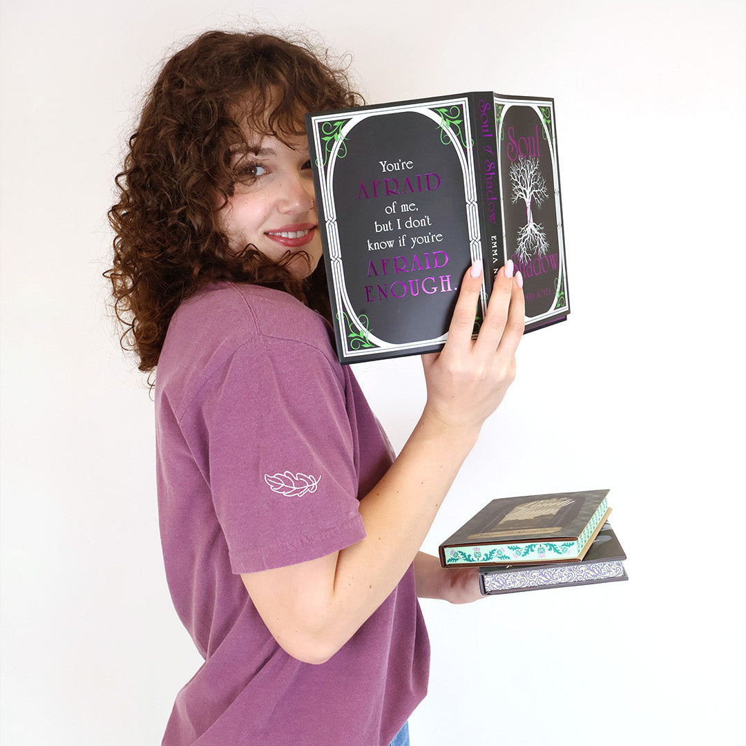 A woman with curly brown hair holds up a book against a plain white background. She is wearing a mauve-purple t-shirt with a white leaf embroidered on the sleeve and blue jeans, and there are two other books stacked in her other hand.